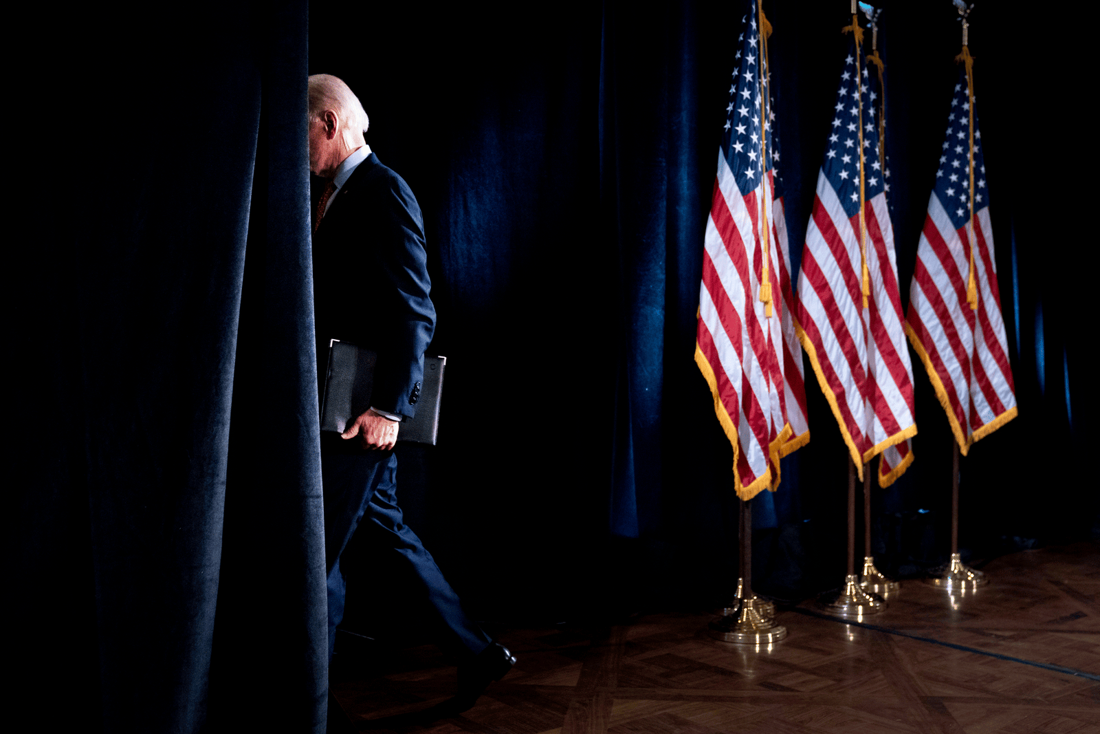 President Joe Biden walking behind a curtain on stage near three United States flags